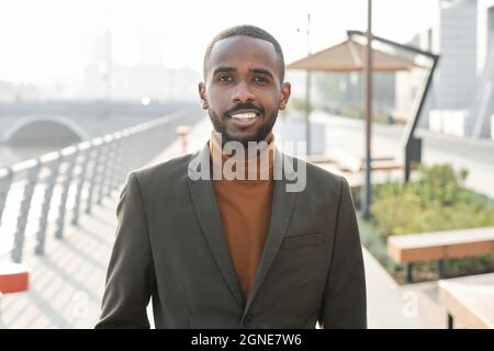 Horizontales mittleres Porträt eines eleganten jungen afroamerikanischen Mannes in eleganter Kleidung, der nach der Arbeit im Büro im Freien steht und lächelt Stockfoto