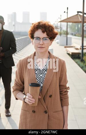 Vertikales mittleres Porträt einer selbstbewussten Frau mit kurzen roten Haaren, die eine im Freien stehende Kaffeetasse hält, nachdem sie im Büro gearbeitet hat und dabei die Kamera betrachtet hat Stockfoto