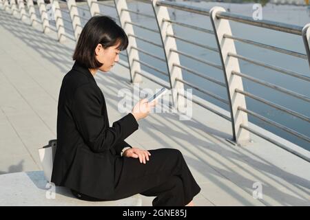 Horizontale Aufnahme einer ernsthaften jungen asiatischen Frau, die modisch elegante Kleidung trägt, die im Freien sitzt und ihren Workflow-Plan auf dem Smartphone überprüft, um Platz zu kopieren Stockfoto