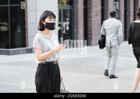 Horizontales mittleres Porträt einer jungen asiatischen Frau, die eine Schutzmaske auf dem Gesicht trägt und das Smartphone im Geschäftsviertel hält, das auf ihren Co-wo wartet Stockfoto