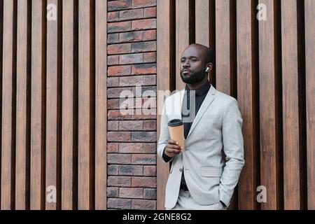 Horizontales mittleres Porträt eines hübschen afroamerikanischen Mannes, der im eleganten grauen Anzug im Freien steht und sich an der Wand lehnt und Musik hört Stockfoto