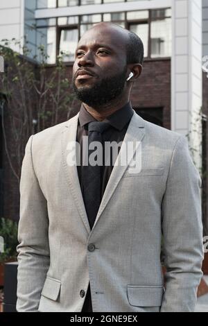 Vertikales mittleres Porträt eines erfolgreichen afroamerikanischen Geschäftsmannes, der Ohrstöpsel trägt, die im Freien stehen und Musik hören, die wegschaut Stockfoto