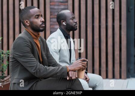 Horizontale, mittlere Seitenansicht von zwei afroamerikanischen Geschäftsleuten, die im Freien auf einer Bank sitzen und ernsthafte Mimik zeigen Stockfoto