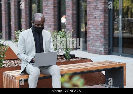 Horizontale Porträtaufnahme eines nicht erkennbaren afroamerikanischen Mannes mit Schutzmaske, der mit einem Laptop auf der Bank im Freien sitzt Stockfoto