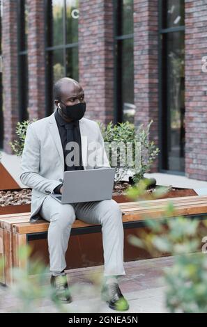 Vertikale Aufnahme eines modernen, nicht erkennbaren afroamerikanischen Mannes mit Schutzmaske auf dem Gesicht, der auf einer Bank im Freien sitzt und auf einem Laptop arbeitet Stockfoto