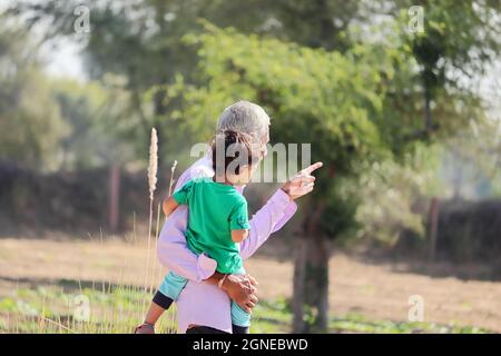 Nahaufnahme eines älteren Großvaters indischer Herkunft, der seinen jüngeren Enkel im Mutterleib hob und mit einer Geste des Ha ein entferntes Objekt auf dem Feld zeigte Stockfoto