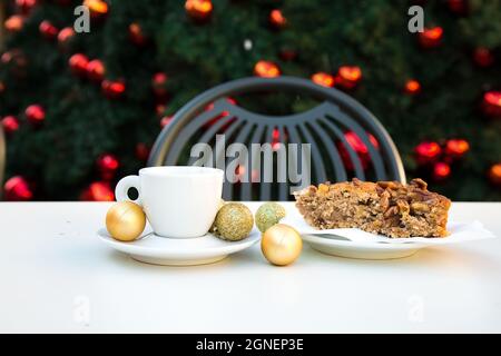Das Kaffeepärchen schließt mit goldenen Weihnachtskugeln und einem Stück Karamellkuchen mit Nüssen. Weißer Tisch, grauer Stuhl und ein Baum mit rotem Spielzeug Stockfoto