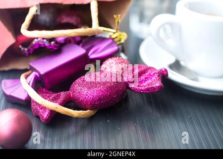 Zwei violette Süßigkeiten Spielzeug für Weihnachtsbaum in der Nähe von rotem Papier Geschenktüte und weißen Kaffeetasse. Nahaufnahme von Neujahr Dekorationen im Café. Winter Urlaub Kaffee Stockfoto