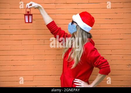 Profil des blonden Mädchens in silberner Maske, rotem Hut und schwarzer Sonnenbrille. Weihnachtslicht in der Hand angehoben. Kaukasische Frau in Weihnachtskostüm und covid Stockfoto