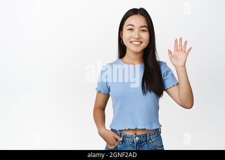 Glückliches asiatisches Mädchen, das hallo sagt, lächelt und winkt mit der Hand, um Sie zu begrüßen und steht in blauem T-Shirt auf weißem Hintergrund Stockfoto