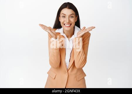 Glückliche Bürofrau im Anzug, spreizte die Hände seitlich und lächelte, demonstrierte das Produkt auf leerem Raum und stand in Firmenkleidung gegen Weiß Stockfoto