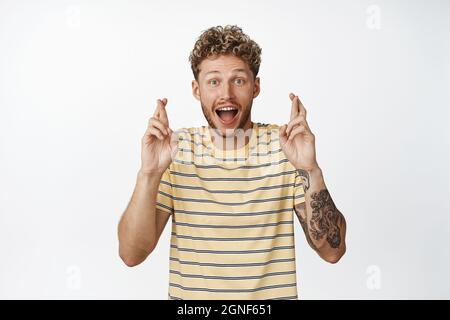 Aufgeregt blonder Mann kreuzt die Finger und schaut hoffnungsvoll, Wünsche machend, wartet auf etwas, vorwegnehmend, stehend in T-Shirt über weißem Hintergrund Stockfoto