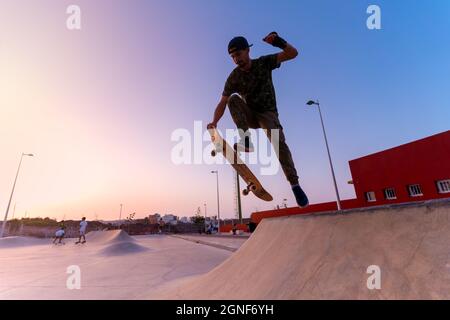 Der junge Skateboarder springt bei Sonnenuntergang mit seinem Board auf die Rampe eines Skateparks. Bewegung 1 Stockfoto