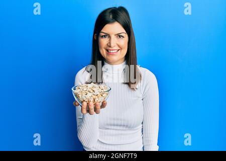 Junge hispanische Frau hält Kürbiskerne Schüssel positiv und glücklich stehend und lächelt mit einem selbstbewussten Lächeln zeigt Zähne Stockfoto