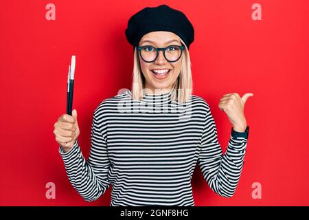 Schöne blonde Frau im französischen Look mit Baskenmütze, die die Pinsel des Malers hält und den Daumen nach oben zur Seite zeigt und glücklich mit offenem Mund lächelt Stockfoto