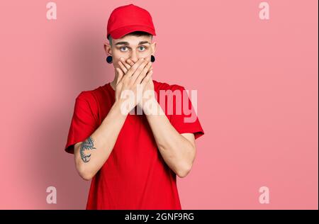 Junge kaukasische Junge mit Ohren Erweiterung tragen Lieferung Uniform und Kappe schockiert decken Mund mit Händen aus Versehen. Secret Konzept. Stockfoto