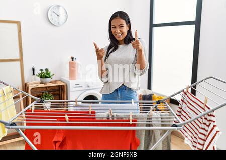 Junge hispanische Frau, die frische Wäsche auf die Wäscheleine legt und mit glücklichen und lustigen Gesichtes die Finger zur Kamera zeigt. Gute Energie und Vibes. Stockfoto