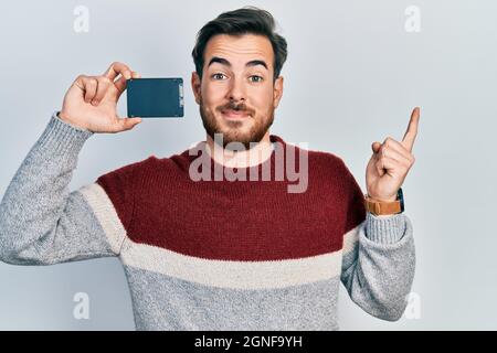 Hübscher kaukasischer Mann mit Bart, der SSD-Speicher hält, lächelt fröhlich und zeigt mit Hand und Finger zur Seite Stockfoto