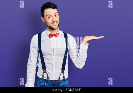 Hispanic Mann mit Bart trägt Hipster-Look mit Fliege und Hosenträger lächelnd fröhlich präsentiert und zeigt mit Handfläche Blick auf die Kamera Stockfoto