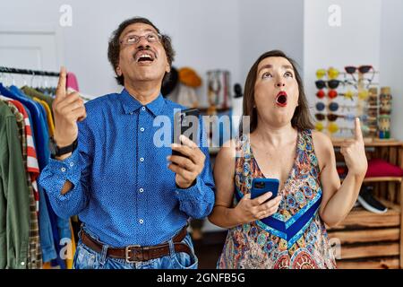 Interrassisches Paar im mittleren Alter im Einzelhandelsgeschäft mit Smartphone erstaunt und überrascht aufschauend und zeigend mit Fingern und erhobenen Armen. Stockfoto