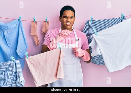 Junger gutaussehender hispanischer Mann mit Reinigungsschürze, der Kleidung auf der Wäscheleine hält, empörte Gesichtsausdruck, unzufrieden und ängstlich, wenn er Ekel macht Stockfoto