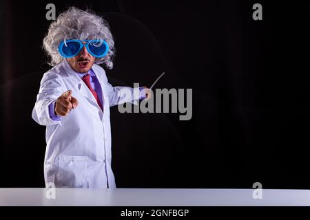 Wütender Wissenschaftslehrer im weißen Mantel mit ungepflegten Haaren in lustigen Augenbrillen zeigt mit seinem Finger auf schwarzen Hintergrund Stockfoto