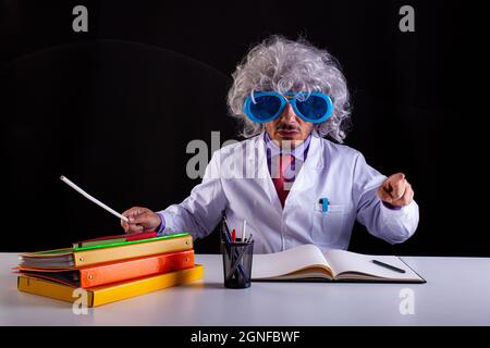 Verrückter Wissenschaftslehrer im weißen Mantel mit ungepflegten Haaren in einer lustigen Augenbrille, die am Schreibtisch sitzt Stockfoto