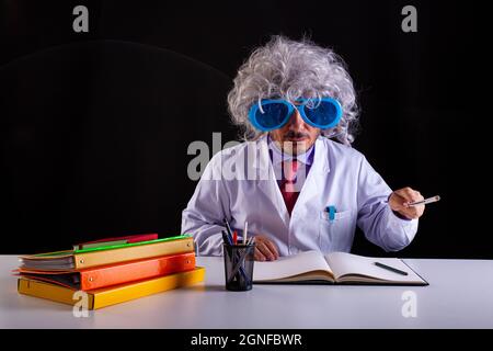 Verrückter Wissenschaftslehrer im weißen Mantel mit ungepflegten Haaren in einer lustigen Augenbrille, die am Schreibtisch sitzt Stockfoto