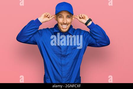 Glatze Mann mit Bart tragen Baumeister Jumpsuit uniform lächelnd ziehen Ohren mit Fingern, lustige Geste. Audition Problem Stockfoto