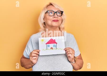 Die blonde Frau aus dem mittleren Alter, die das Haus hält, schaut lächelnd zur Seite und starrt denkend weg. Stockfoto