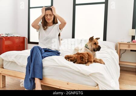 Junge Brünette Frau sitzt auf dem Bett mit zwei Hunden leiden unter Kopfschmerzen verzweifelt und gestresst, weil Schmerzen und Migräne. Hände auf dem Kopf. Stockfoto