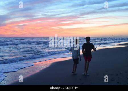 Rückansicht von zwei Jungen im Teenageralter, die bei Sonnenuntergang am Ufer des Sandstrandes mit Angelruten spazieren, Marina di Castagneto Carducci, Toskana Stockfoto