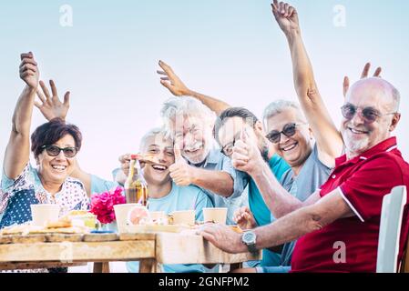 Porträt einer fröhlichen Familie mit mehreren Generationen, die das Essen und Trinken am Tisch im Freien zelebriert. Aufgeregtes Familienjubeln, das Daumen nach oben gibt Stockfoto