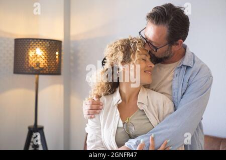 Paar umarmt und romantisch stehend im Wohnzimmer zu Hause. Ehemann küsst die Stirn der Frau mit Zuneigung. Liebevolles Paar, das Freizeit verbringt Stockfoto