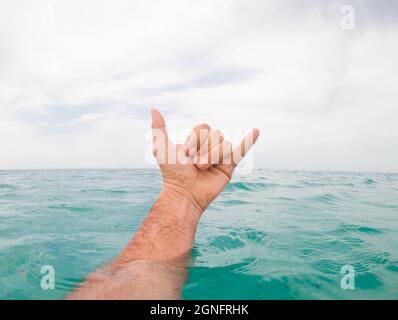 Nahaufnahme der Hand eines Mannes, der das shaka-Schild über dem Meerwasser gegen den Himmel zeigt. Hand eines Mannes, der aus der Meerwasseroberfläche kommt und shaka Zeichen gegen gestikuliert Stockfoto