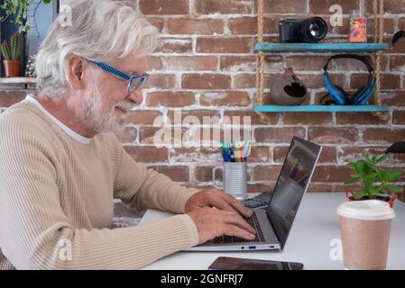Profil eines älteren Mannes, der mit einem Laptop auf dem Schreibtisch und einer Einweg-Kaffeetasse Social-Media-Inhalte durchsucht. Alter pensionierter Mann, der seine Zeit damit verbringt, an ihm zu arbeiten Stockfoto