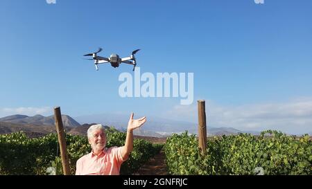 Drohne fliegt über das Feld, wobei ein älterer Mann die Hand hebt, um sie gegen Berg und Himmel zu fangen. Älterer Mann, der Drohne über Ackerland benutzt. Alter Mann mit Fliegen Stockfoto