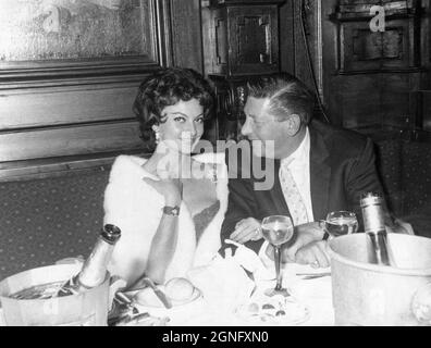 Eine elegante und attraktive junge Frau, die eine Pelzstola trägt, genießt ein Essen mit einem älteren Herrn im Society Restaurant, Jermyn Street, London. Anfang der 1960er Jahre. Die Dame lächelt der Kamera zu, während der Mann sie anschaut. Auf dem Tisch befinden sich Flaschen Champagner, die in Eiskübeln kühlen, und Petit Fours auf einem Teller davor. Stockfoto