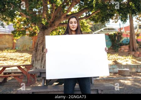 Teenager-Aktivist mit einem leeren Plakat im Freien in der Stadt. Selbstbewusstes Teenager-Mädchen, das die Kamera anschaut, während es ein weißes Banner in einem städtischen p hält Stockfoto