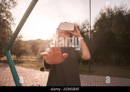 Nahaufnahme eines Mädchens, das mit den Händen eine Virtual-Reality-Brille auf dem Kopf hält und auf einem Hintergrund von grünen Bäumen steht. Er reicht eine Hand an die Kamera. Hochwertige Fotos Stockfoto