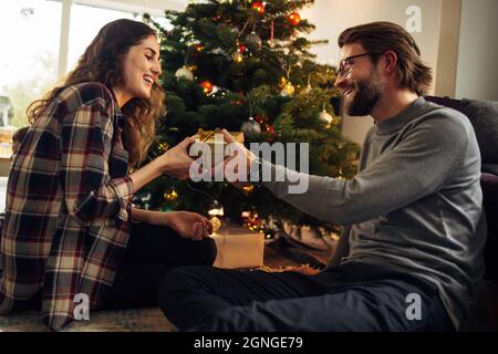 Mann, der seiner Frau zu Hause ein Geschenk macht. Paar sitzt am Weihnachtsbaum und tauscht Geschenke aus und lächelt. Stockfoto