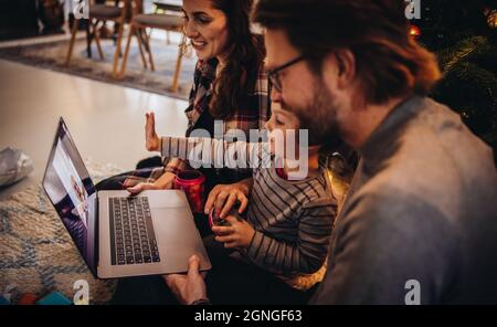 Familie, die während der weihnachtszeit auf dem Boden sitzt und einen Videoanruf auf dem Laptop führt. Familie begrüßt Freunde während eines Videoanrufs. Stockfoto