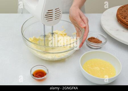 Den Schokoladenkuchen zum Zuckerguss machen. Frau Hände schlagen Butter in einer Glasschüssel mit einem elektrischen Handmixer, Nahaufnahme Stockfoto