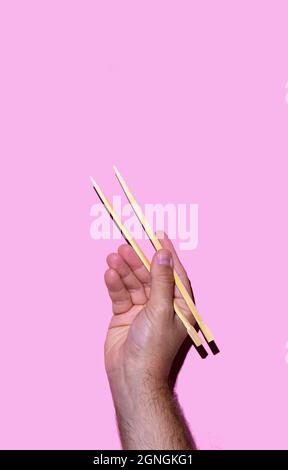 Hand eines Mannes, der japanische Essstäbchen auf rosa Hintergrund hält. Sushi- und japanisches Speisekonzept Stockfoto