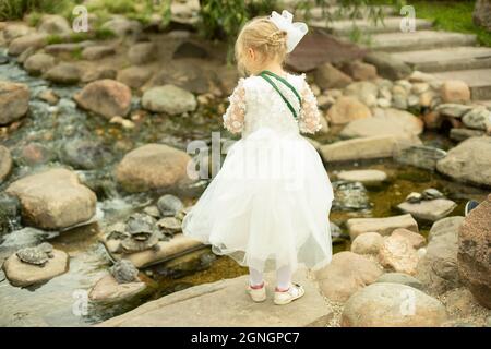 Vorschulkinder in einem weißen Kleid. Kind im Park. Das Mädchen schaut auf den Strom. Die Brautjungfer geht. Kind auf der Party. Stockfoto