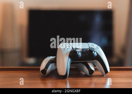 London, Großbritannien – 25. Mai 2021: Zwei PlayStation 5 (PS5)-Controller auf dem Tisch, tv im Hintergrund, selektiver Fokus. PlayStation 5 ist das neueste Videospiel Stockfoto