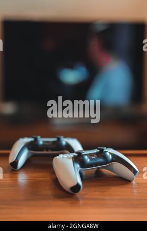 London, Großbritannien – 25. Mai 2021: Nahaufnahme der PlayStation 5 (PS5)-Controller auf dem Tisch, tv im Hintergrund, selektiver Fokus. PlayStation 5 ist das neueste vi Stockfoto
