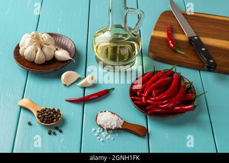 Gewürzset auf einem rustikalen, türkisfarbenen Holztisch. Rohe rote Chili-Paprika, Knoblauchkopf und Nelken, Meersalz, schwarze Pfefferkörner und ein Glas Olivenöl. Stockfoto