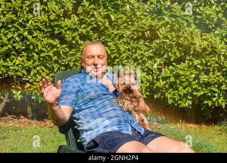 Reifer Mann mit Yorkshire Terrier Hund unter Spritzwasser an heißen Tag im Garten. Stockfoto