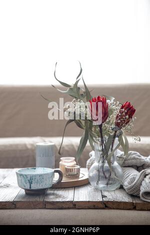 Tasse, Kerzen, Vase mit protea-Blumen und einem gestrickten Element im Raum auf verschwommenem Hintergrund. Stockfoto